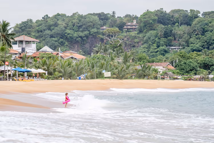 Kind mit rosa Schwimmring in der Brandung an einem Sandstrand in Sri Lanka, dahinter Palmen und ein bewaldeter Hang