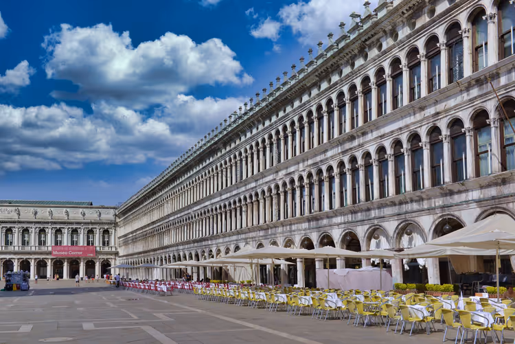Lunga facciata porticata delle Procuratie in Piazza San Marco a Venezia, con sedie gialle dei caffè e ombrelloni