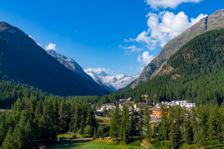 Kleines Dorf im Lärchenwald in einem Schweizer Alpental, in der Ferne schneebedeckte Gipfel und ein Gletscher