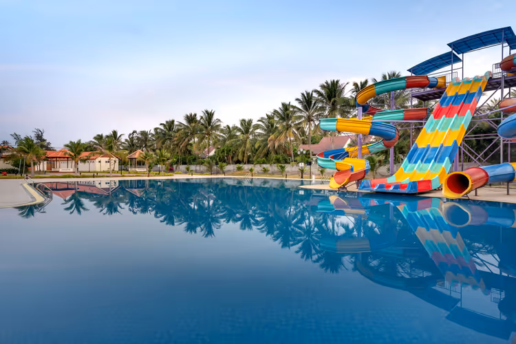 Bunt gestreifte Wasserrutschen neben einem ruhigen blauen Pool, in dem sich eine Palmenreihe unter blassem Himmel spiegelt