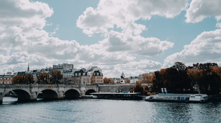 Steinerne Bogenbrücke über die Seine in Paris mit vertäuten Flussschiffen, Herbstbäumen und Dächern unter bewölktem Himmel