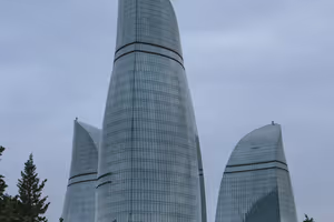 Die drei geschwungenen gläsernen Flame Towers in Baku vor grauem, bedecktem Himmel, im Vordergrund Tannenwipfel