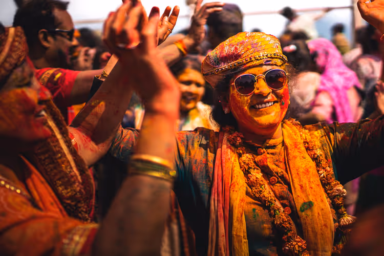 Lächelnde Frau mit Sonnenbrille und Turban, voller Farbpulver, tanzt in der Menge beim Holi-Fest in Indien