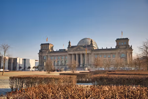 Reichstagsgebäude in Berlin mit Glaskuppel und Deutschlandfahnen unter klarem blauem Himmel, davor kahle Winterhecken