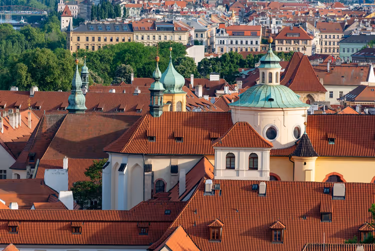 Rote Ziegeldächer von Prag mit grünen Kupferkuppeln und barocken Kirchtürmen, von oben zwischen Bäumen gesehen