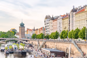 Moldauufer in Prag mit festgemachten Booten, Spaziergängern, einem steinernen Wasserturm und verzierten Stadthäusern