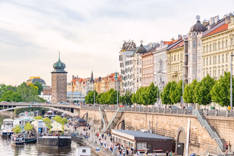 Moldauufer in Prag mit festgemachten Booten, Spaziergängern, einem steinernen Wasserturm und verzierten Stadthäusern
