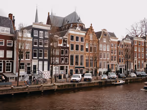 Schmale Giebelhäuser an einer Gracht in Amsterdam mit geparkten Transportern, Fahrrädern und einem kleinen Boot unter grauem Himmel