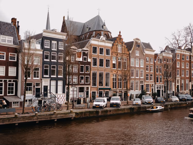 Schmale Giebelhäuser an einer Gracht in Amsterdam mit geparkten Transportern, Fahrrädern und einem kleinen Boot unter grauem Himmel