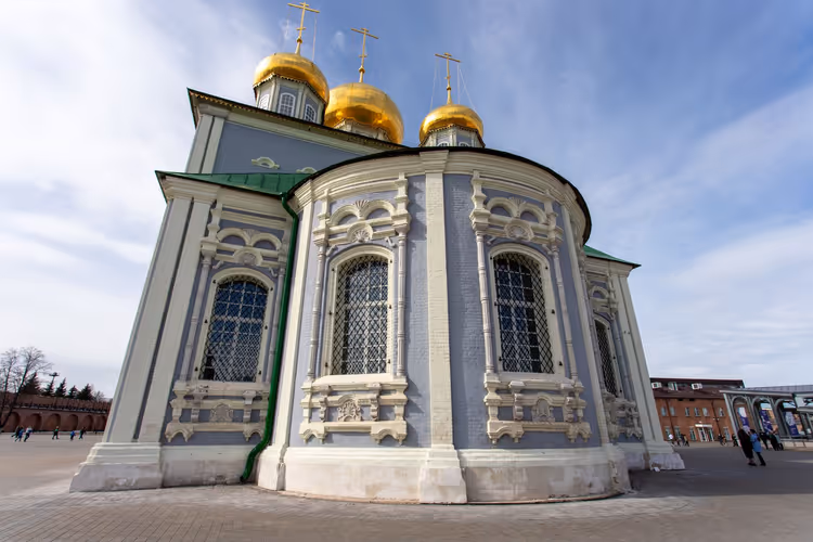 Hellblaue orthodoxe Kirche mit weißen barocken Fensterrahmen und drei goldenen Kuppeln, von unten auf einem gepflasterten Platz gesehen