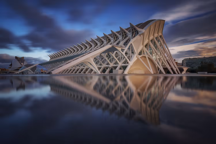 Weißes, geripptes Gebäude des Wissenschaftsmuseums in Valencia spiegelt sich in der Dämmerung unter dunklen Wolken in einem stillen Becken