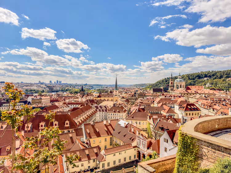 Rote Ziegeldächer der Prager Kleinseite von einer Hangterrasse aus, mit Kirchenkuppel und bewaldetem Hang