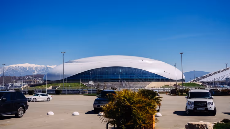 Weiße Eisarena mit Kuppel in Sotschi unter klarem blauem Himmel, dahinter schneebedeckte Berge, davor eine Palme auf dem Parkplatz