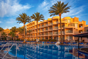Oranges Resorthotel mit geschwungenen Balkonen und hohen Palmen spiegelt sich bei Sonnenuntergang in einem großen Pool in Hurghada
