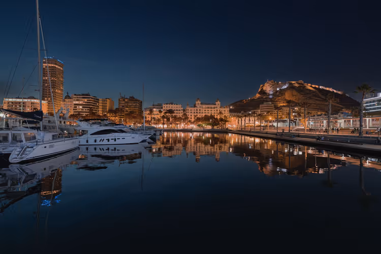 Yachthafen von Alicante bei Nacht, Yachten und beleuchtete Uferhäuser spiegeln sich im stillen Wasser unterhalb des Burgbergs