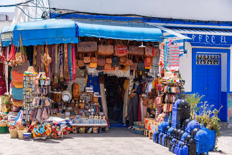 Souvenirstand mit Ledertaschen, Flechtkörben, Textilien und blauen Laternen unter einer blauen Markise neben einer blauen Tür