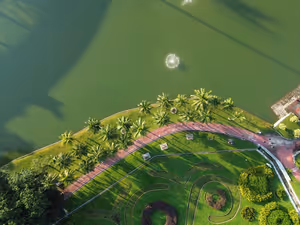 Luftaufnahme eines Parks am See in Malaysia mit Palmen, geschwungenem rosa Weg und Springbrunnen im grünen Wasser