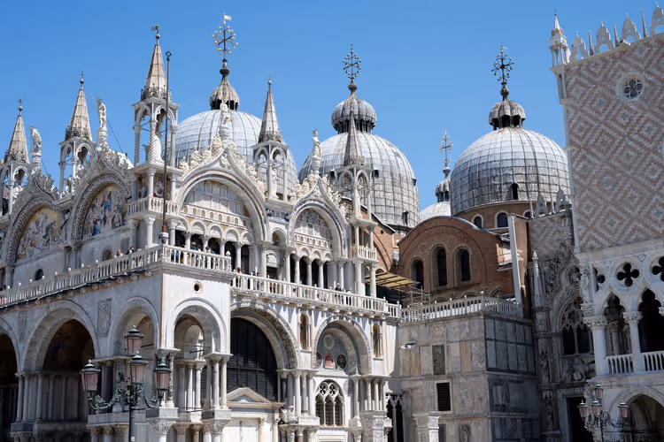 Kuppeln, Turmspitzen und weiße Marmorbögen mit Mosaiken an der Fassade des Markusdoms in Venedig unter blauem Himmel