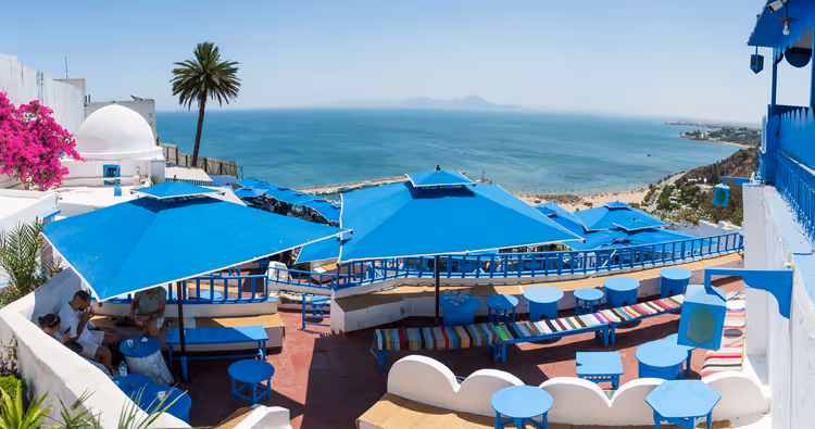 Caféterrasse mit leuchtend blauen Schirmen und Tischen über einer türkisfarbenen Bucht, weißer Kuppelbau und Palme in Tunesien