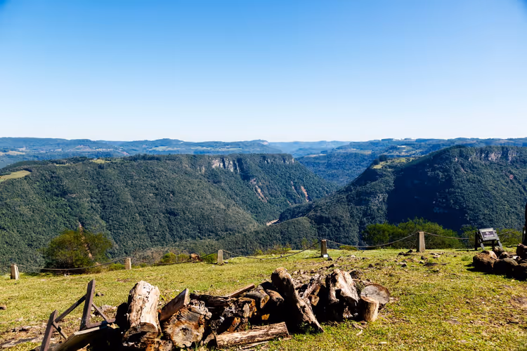 Stapel geschnittener Baumstämme auf einer grasbewachsenen Hügelkuppe mit Blick auf bewaldete Schluchten und Bergrücken unter blauem Himmel