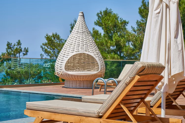 Holzliegen mit beigen Auflagen und ein weißes Kokon-Daybed aus Korbgeflecht auf einem Pooldeck inmitten von Kiefern