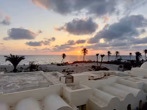 Sonnenuntergang über dem Meer in Tunesien, gesehen über weiße Kuppeldächer und eine Palmenreihe an einem Sandstrand hinweg