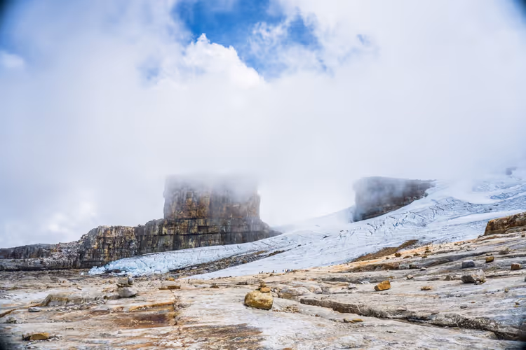 Wolkenverhangene Felstürme mit flachen Gipfeln über einem Schneefeld und kahlen Steinplatten auf einer Hochebene