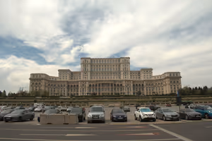 Gewaltiger Parlamentspalast in Bukarest unter bewölktem Himmel, im Vordergrund ein Parkplatz voller Autos