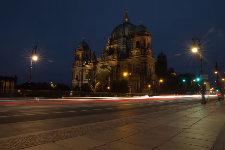 Berliner Dom mit seinen Kuppeln bei Nacht, leuchtende Straßenlaternen und Lichtspuren vorbeifahrender Autos auf der Straße davor