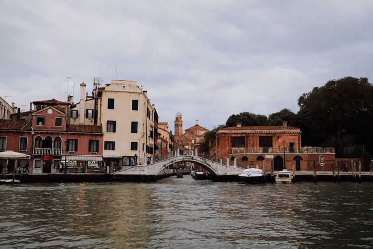 Piccolo ponte ad arco e case rosse e color crema segnate dal tempo lungo un canale di Venezia sotto un cielo coperto