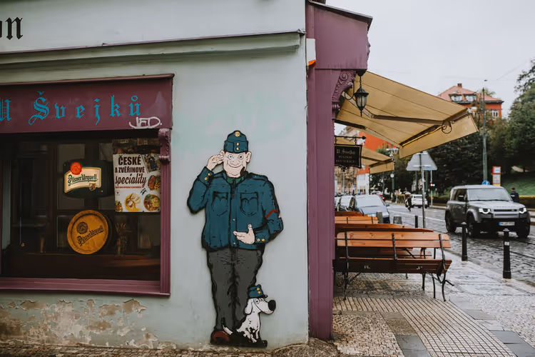 Bemalte Figur eines Soldaten mit Hund an der Ecke einer Prager Kneipe, Holzbänke unter Markisen an einer regennassen Straße