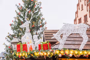 Weiße Rentiere aus Draht und rote Geschenkpakete auf dem Dach einer Weihnachtsmarktbude, dahinter eine geschmückte Tanne