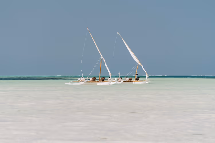 Zwei traditionelle Dau-Segelboote mit schrägen Masten liegen im flachen türkisfarbenen Wasser vor Sansibar
