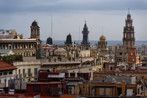 Dächer, Kuppeln und verzierte Türme der Altstadt von Barcelona, am Horizont das Meer unter grauem Himmel