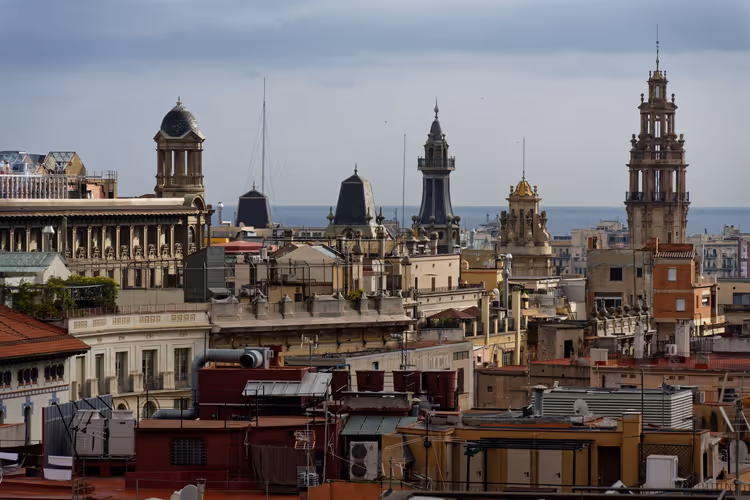 Dächer, Kuppeln und verzierte Türme der Altstadt von Barcelona, am Horizont das Meer unter grauem Himmel