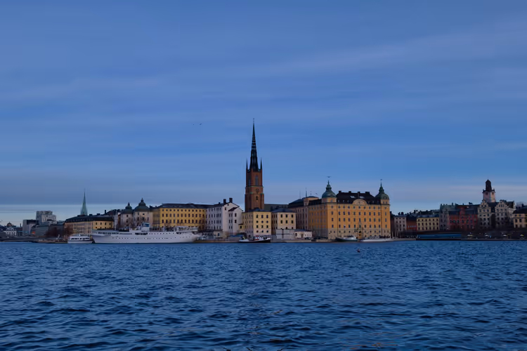 Uferpromenade in Stockholm in der Abenddämmerung, eine dunkle Kirchturmspitze ragt über gelben Gebäuden jenseits des blauen Wassers auf