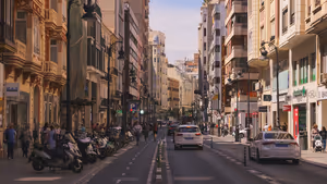 Sonnige Straße in Valencia mit verzierten Balkonfassaden, geparkten Motorrollern, Autos und Fußgängern