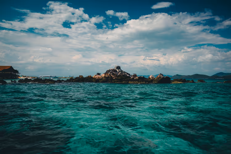 Türkises, leicht gekräuseltes Meer mit kleiner Felseninsel und Pfahlbau links unter weißen Wolken in Thailand