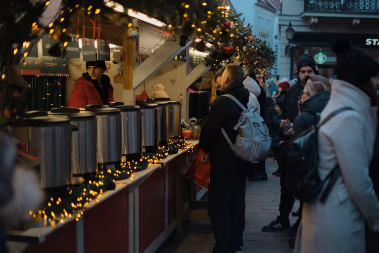 Besucher in Wintermänteln an einem Weihnachtsmarktstand mit Lichterketten und großen Getränkekesseln aus Metall in der Dämmerung