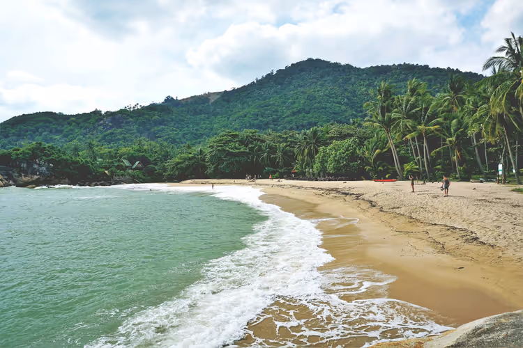 Sandstrand auf Koh Samui mit schäumender Brandung, grünem Wasser, Kokospalmen und einem dschungelbedeckten Hügel unter Wolken