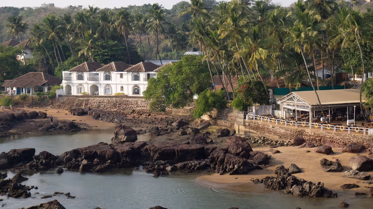 Weißes Haus im Kolonialstil und Strandcafé zwischen Kokospalmen über einem felsigen Sandufer in Goa