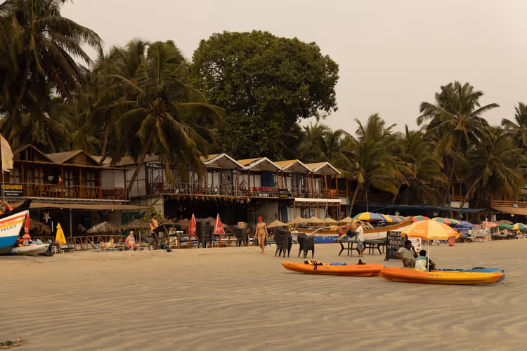 Sandstrand in Goa mit orangefarbenen Kajaks, bunten Sonnenschirmen, Kühen und hölzernen Strandhütten unter hohen Palmen