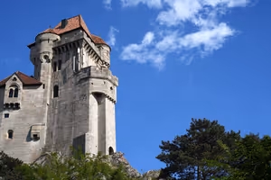 Heller steinerner Burgturm mit rotem Ziegeldach auf einem Felsen über grünen Bäumen vor tiefblauem Himmel