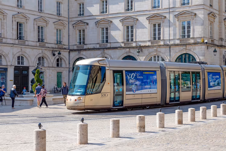 Moderne beige Straßenbahn vor einem klassizistischen Steingebäude auf einem gepflasterten autofreien Platz mit Fußgängern und Pollern