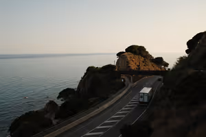 Lkw auf einer kurvenreichen Küstenstraße oberhalb eines ruhigen Meeres, felsige Landzungen im weichen Dämmerlicht