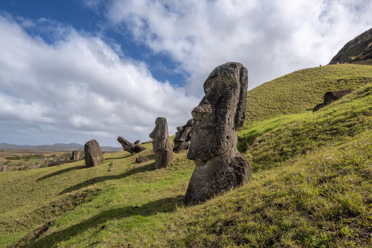Halb im grasbewachsenen Hang vergrabene Moai-Steinköpfe auf der Osterinsel, dahinter weitere schief stehende unter bewölktem Himmel
