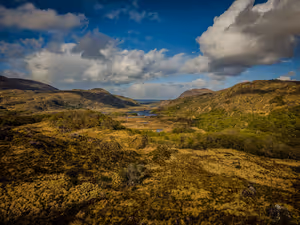 Weites Tal mit Seen zwischen schroffen Hügeln unter großen Wolken, Moos und Heidekraut im Killarney-Nationalpark