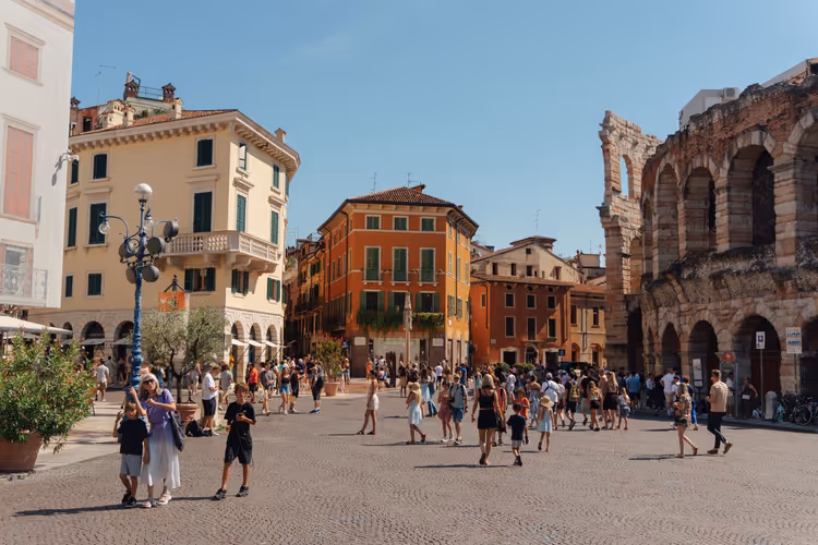 Viele Menschen überqueren einen sonnigen Pflasterplatz in Verona neben den Bögen der römischen Arena und pastellfarbenen Stadthäusern