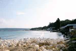 Weiße Felsblöcke am Ufer, Schwimmer im ruhigen Meer, dahinter ein Sandstrand und bewaldete Hügel im Bogen