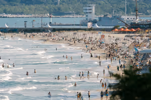 Voller Sandstrand mit Badenden in der Brandung, Reihen von Sonnenschirmen und im Hintergrund ein Hafen mit Schiffen